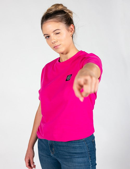 A woman wearing a bright pink short-sleeve crewneck t-shirt with a small black Rocket Science chest logo, paired with blue jeans, pointing towards the camera against a light gray background.