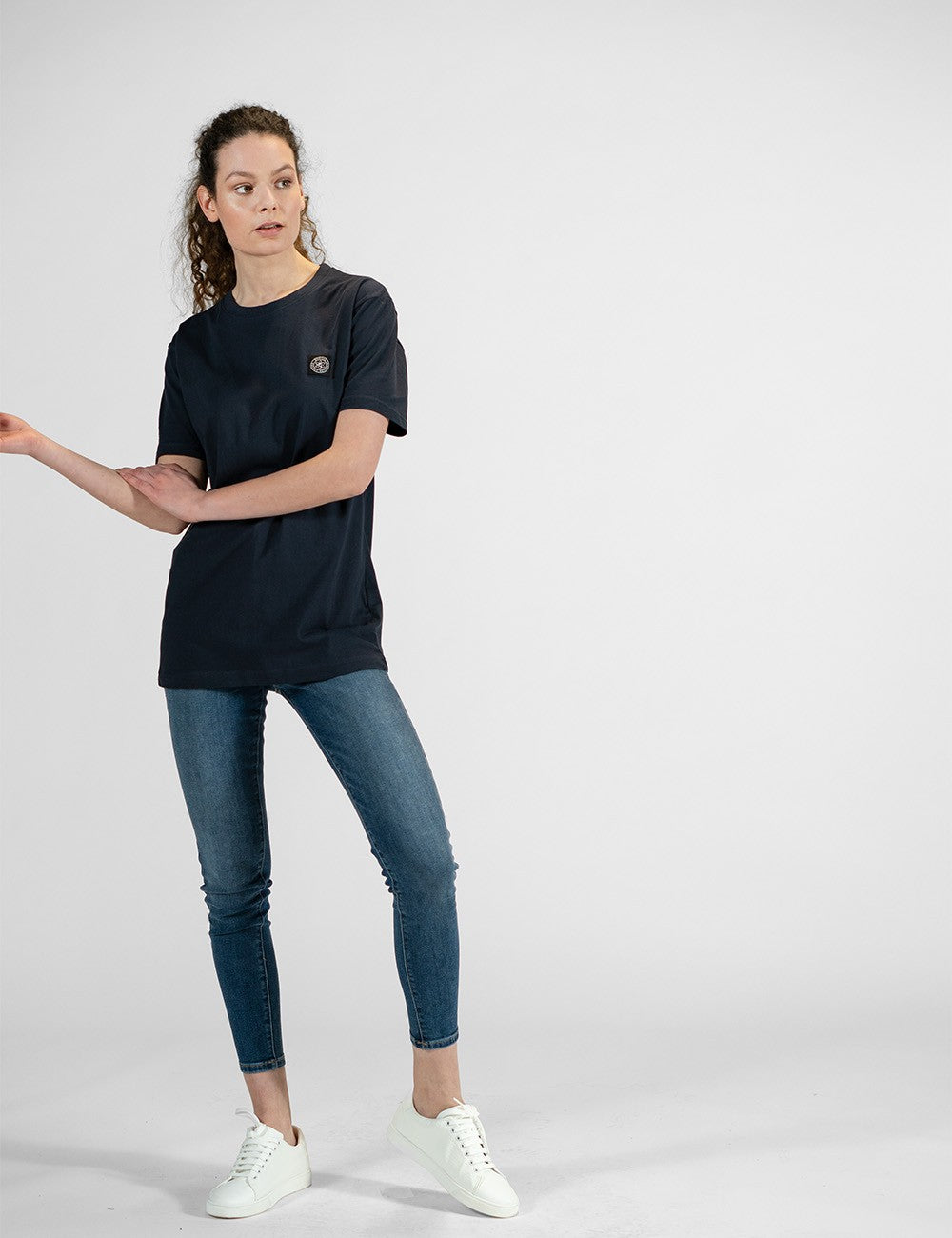This image shows a young woman wearing a navy blue round-neck T-shirt, blue slim-fit jeans, and white sneakers.