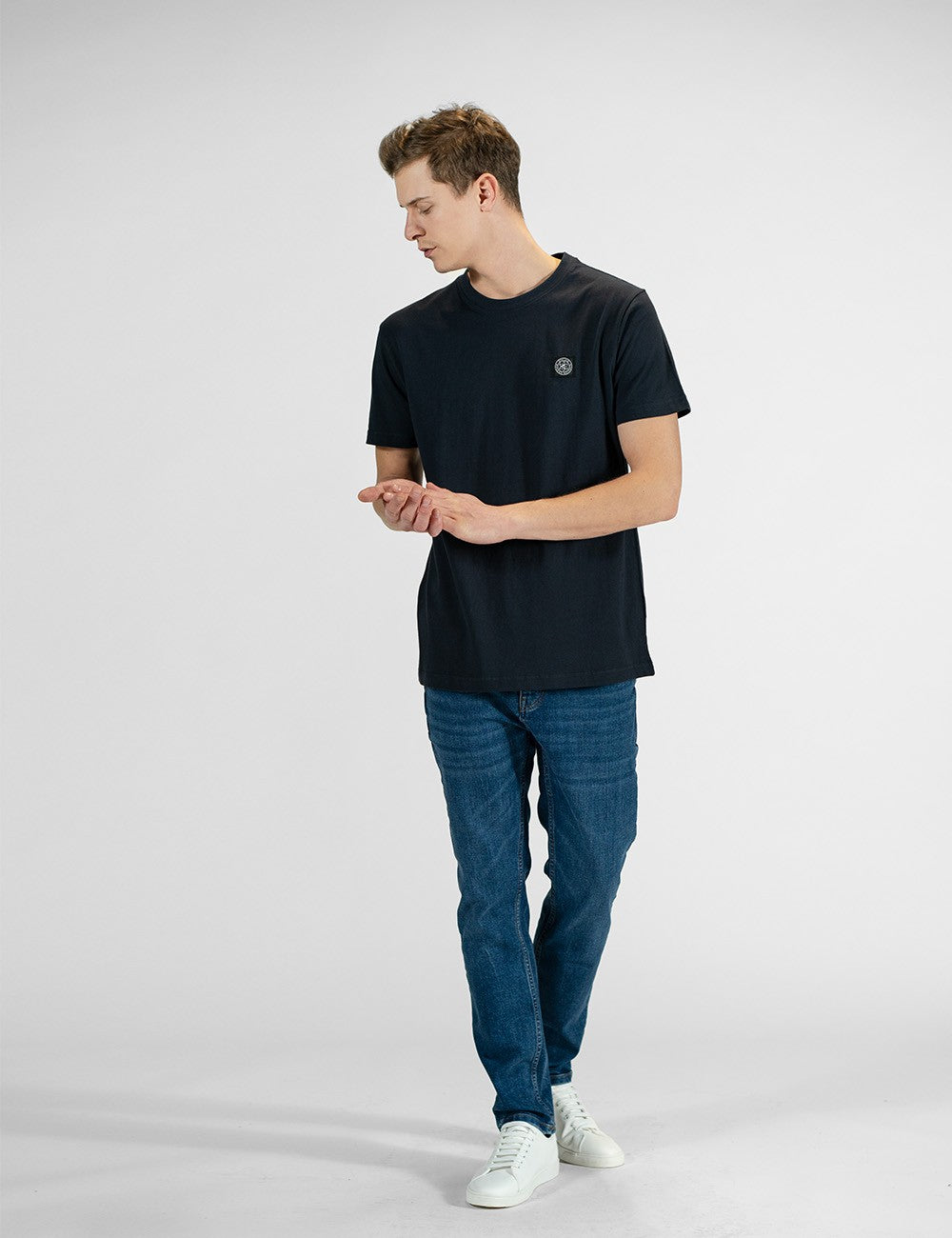 A young man with light brown hair stands looking down and to his right, wearing a classic navy blue crew neck t-shirt with a small circular logo on the left chest, medium-wash blue jeans, and clean white sneakers.