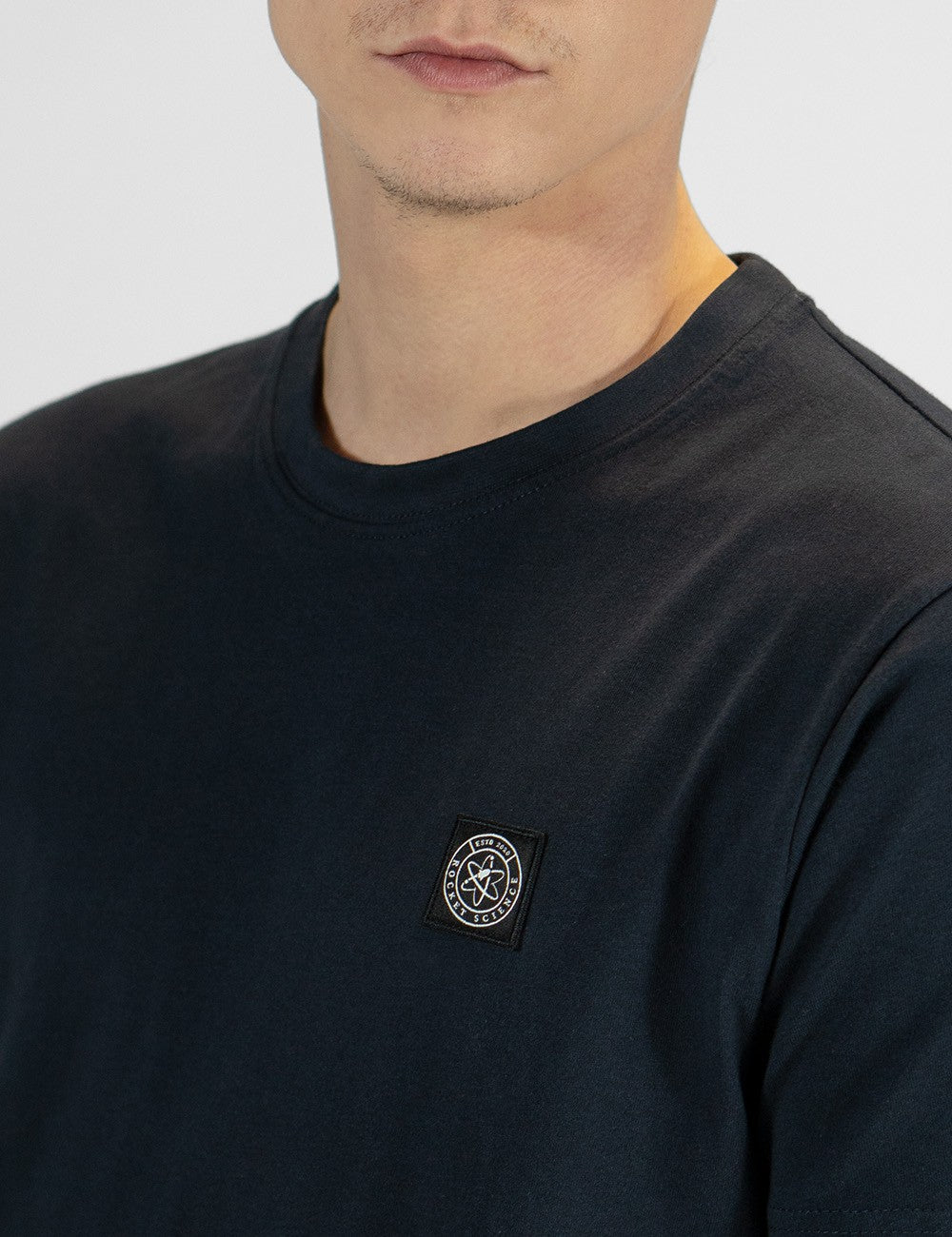 The image shows a dark crew-neck cotton t-shirt featuring a small round logo patch on the chest.