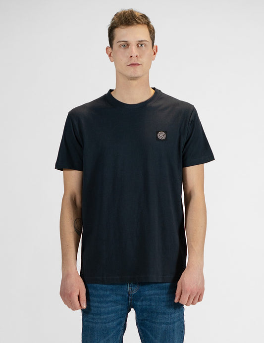 A young man with light brown hair stands against a plain white background, wearing a navy blue crewneck T-shirt with a small circular logo patch on the left chest, paired with blue jeans.