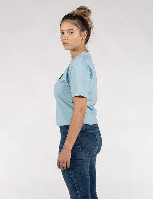 A woman in a light blue short-sleeve crop top and dark blue jeans, shown from a side profile against a plain light gray studio background.