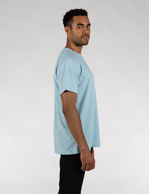 A man in profile facing right, wearing a light blue crew-neck t-shirt and black pants, against a plain white background.