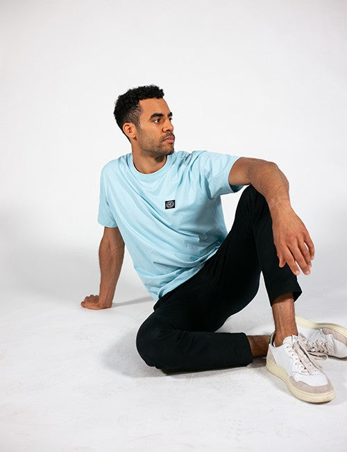 A man sitting on a white studio floor wearing a light blue crewneck t-shirt with a small black square logo on the left chest, black pants, and white sneakers.