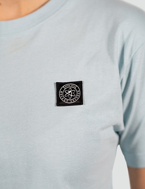 A close-up shot focusing on the chest area of a light blue t-shirt. On the left side of the chest is a small, square black fabric patch. The patch features a white embroidered circular logo containing an atom symbol in the center, surrounded by the text "ROCKET SCIENCE".
