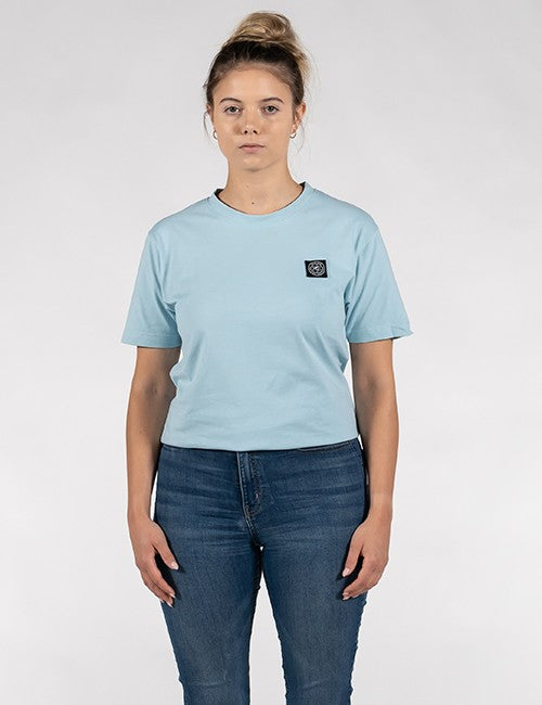 A woman modeling a light blue short-sleeve crewneck t-shirt with a small black square logo patch on the left chest, paired with dark blue jeans, against a light gray studio background.