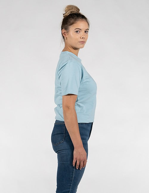 A woman wears a light blue crew-neck short-sleeve t-shirt and dark blue skinny jeans, set against a plain light gray background.