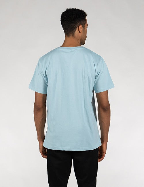 A man viewed from behind wearing a light blue crewneck t-shirt and black pants, standing against a neutral light gray studio background.