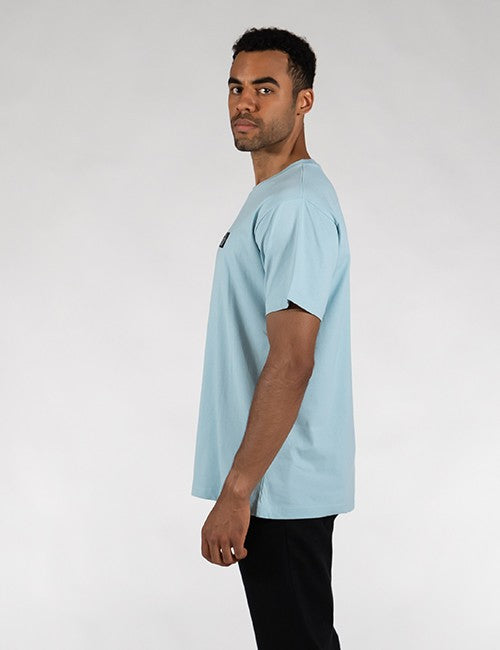 A man wearing a light blue crew-neck t-shirt and black pants, shown in profile against a plain light gray studio background.