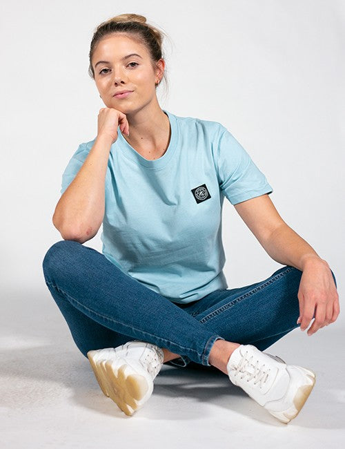 A woman sitting cross-legged on a light gray studio floor wearing a light blue crewneck t-shirt with a small black square logo patch on the left chest, blue jeans, and white sneakers.