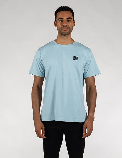 A man wearing a light blue crewneck t-shirt with a small black square logo patch on the left chest, paired with black pants, standing against a light gray studio background.