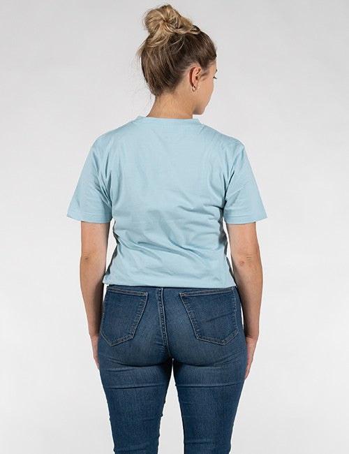 A woman viewed from behind wearing a light blue short-sleeve t-shirt and dark blue jeans, standing against a plain light gray studio background.