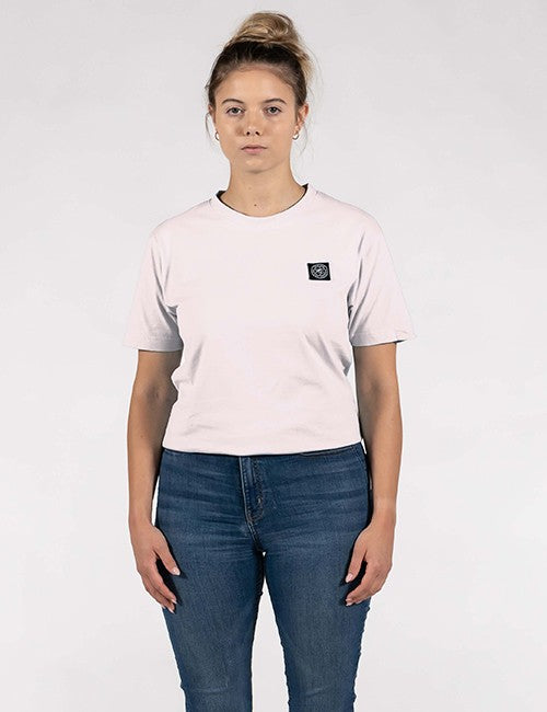 A woman modeling a light pink short-sleeve crewneck t-shirt with a small black square logo patch on the left chest, paired with blue jeans, against a plain light gray background.