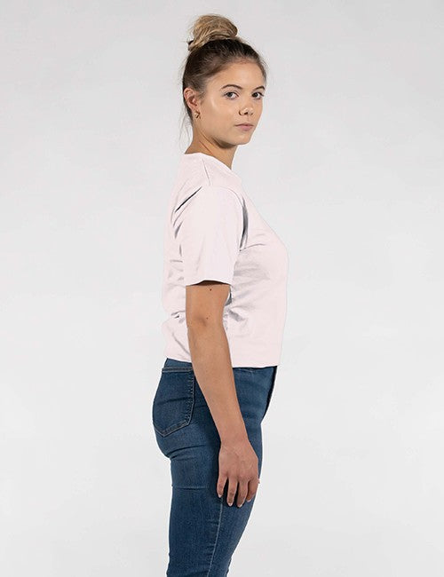 A woman shown in side profile wearing a light pink short-sleeve crop top and dark blue jeans against a plain light gray studio background.