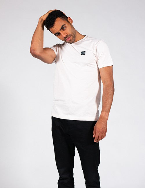 A man wearing a white crew-neck t-shirt with a small black square logo patch on the left chest, paired with black pants, posing against a light gray studio background.