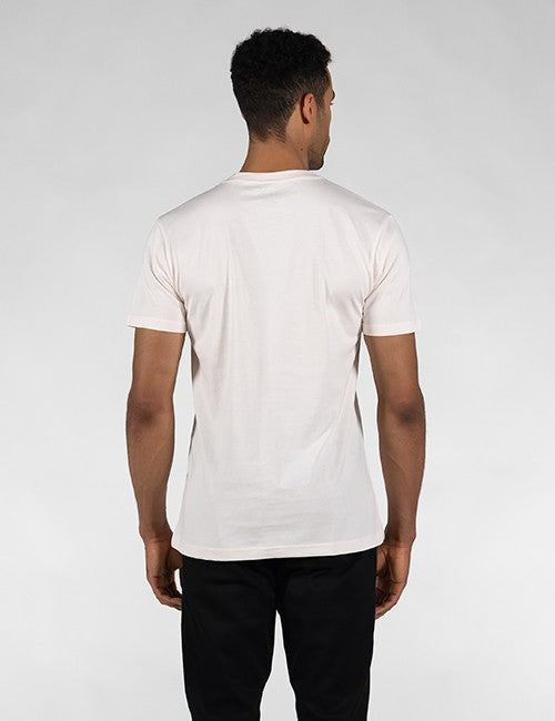 A man facing away from the camera, wearing a light beige or off-white short-sleeve crew-neck T-shirt and black pants against a plain, light grey background.