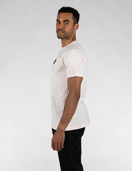 A side-profile shot of a man standing against a plain grey background, wearing a light pink, short-sleeved crewneck t-shirt with a small black graphic on the front left chest, paired with black trousers.