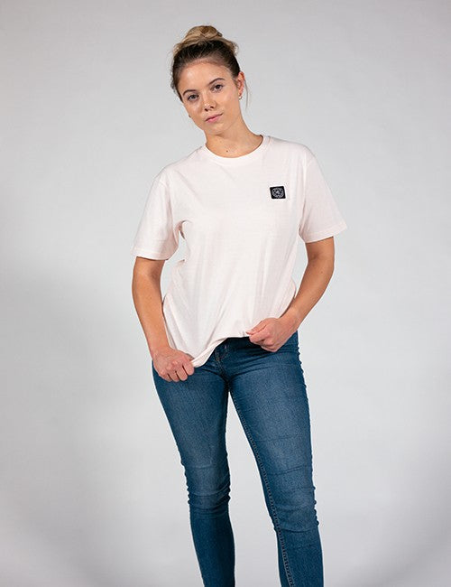 A woman modeling a light pink short-sleeve t-shirt with a small black square logo patch on the left chest, paired with blue jeans, against a light gray studio background.