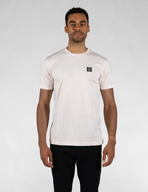 A man with short dark hair and a light beard stands facing forward against a plain gray background. He is wearing a light pink, short-sleeved round-neck t-shirt with a small, square black logo patch on the left chest, paired with black pants.