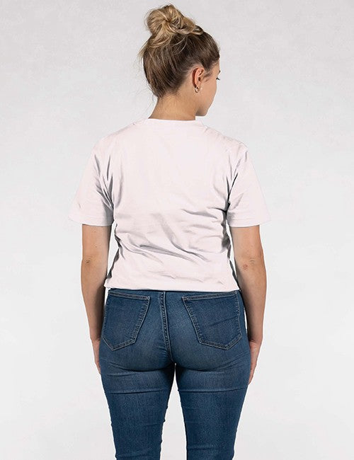 A woman seen from behind is wearing a white or light pink, short-sleeved crewneck t-shirt tucked slightly into dark blue, mid-to-high-waisted denim jeans. Her blonde hair is styled up into a casual high bun, and she stands against a plain, light grey background.