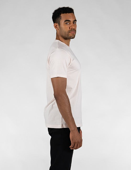 A side-profile shot of a man, wearing a light pink, short-sleeved t-shirt and slim-fit black jeans, looking over her shoulder toward the camera against a plain white background.