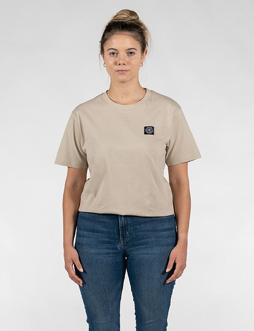 A woman modeling a beige short-sleeve crewneck t-shirt with a small black square Rocket Science logo patch on the left chest, paired with blue jeans, against a light gray studio background.