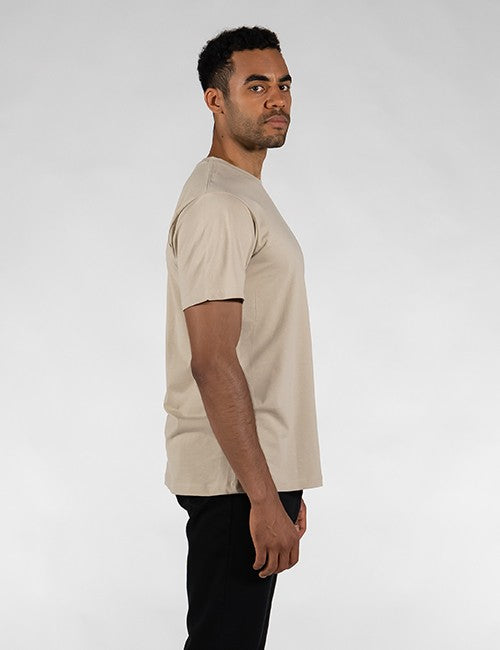 A man in a side profile wearing a light beige short-sleeve crewneck t-shirt and black pants, photographed against a neutral light gray studio background.