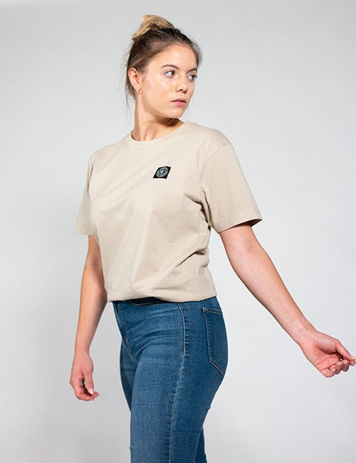 A woman wearing a beige short-sleeve t-shirt with a small black square Rocket Science logo on the left chest, paired with blue jeans, photographed against a light gray studio background.