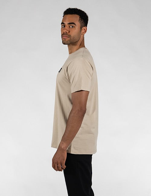 A man wearing a light beige crew-neck t-shirt and black pants, shown in profile against a light gray studio background.