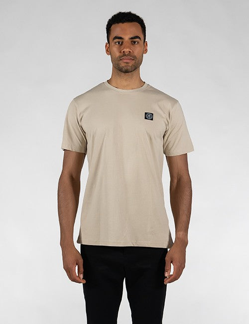 A man wearing a light beige crew-neck t-shirt with a small black square Rocket Science logo patch on the left chest, paired with black pants, standing against a light gray studio background.