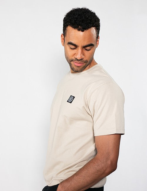 A man wearing a light beige crewneck t-shirt with a small black square Rocket Science logo patch on the left chest, photographed against a plain white studio background.