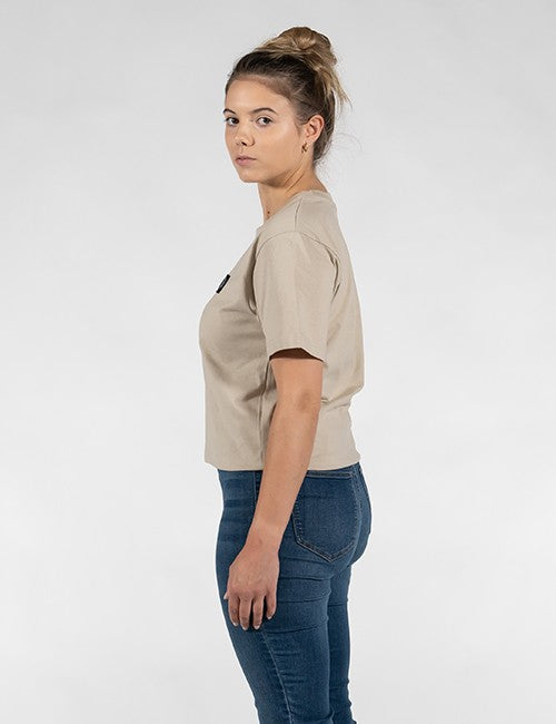 A woman shown in profile wearing a light beige short-sleeve crop top and dark blue jeans against a plain light gray studio background.