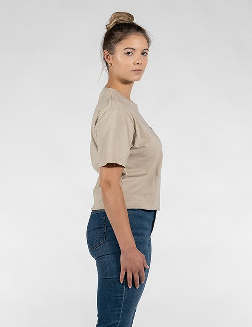 A woman shown in side profile wearing a light beige short-sleeve cropped t-shirt and dark blue jeans, standing against a plain light gray studio background.