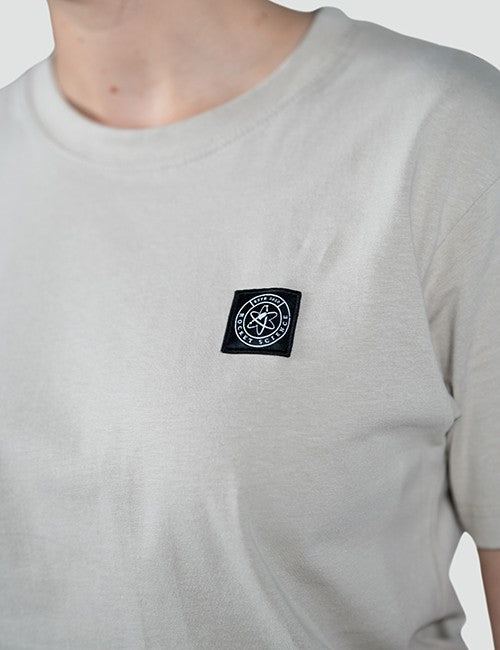 A close-up of a person wearing a light gray crew-neck t-shirt with a black square patch featuring the Rocket Science logo on the left chest.