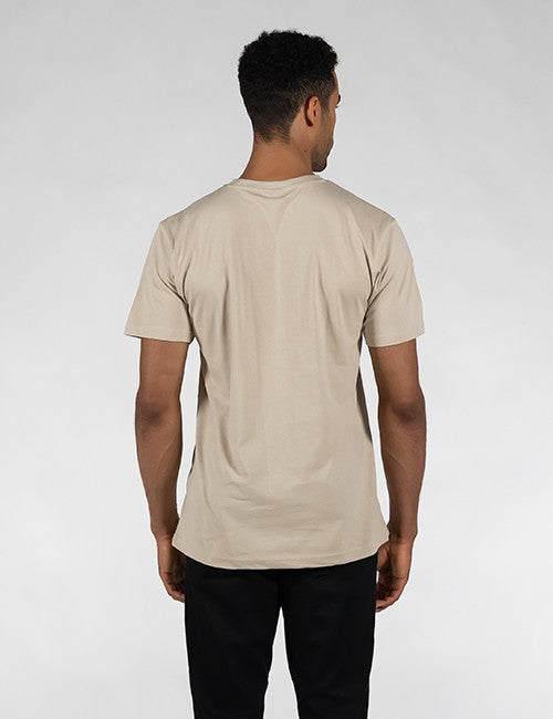 A man viewed from behind wearing a light beige short-sleeve crewneck t-shirt and black pants, standing against a light gray studio background.