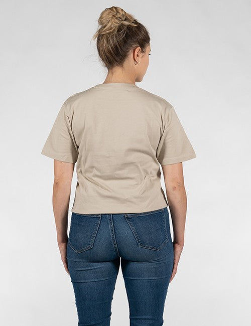 A woman viewed from behind wearing a light beige short-sleeve t-shirt and blue jeans against a plain light gray background.