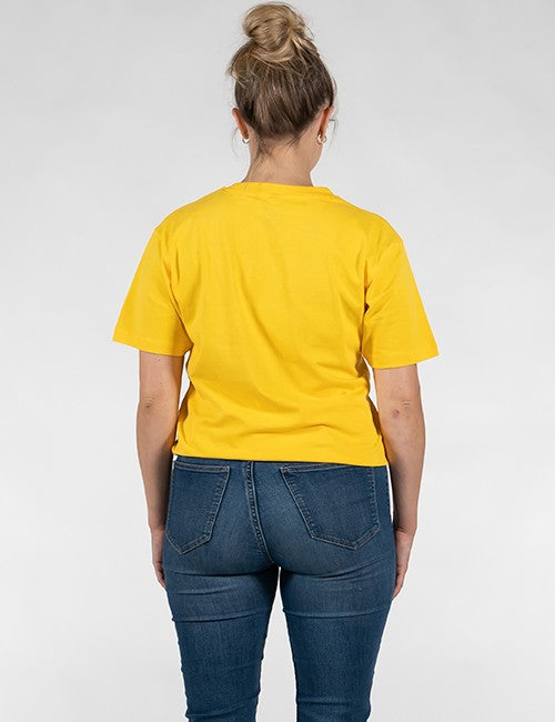 A woman viewed from behind wearing a bright yellow short-sleeve t-shirt and dark blue jeans against a light gray studio background.