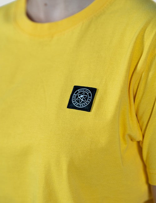 A close-up of a person wearing a bright yellow crewneck t-shirt with a black square Rocket Science logo patch on the left chest.