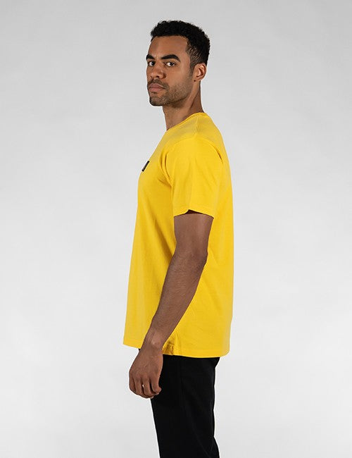 A man wearing a bright yellow short-sleeve crewneck t-shirt and black pants, shown in profile against a light gray studio background.