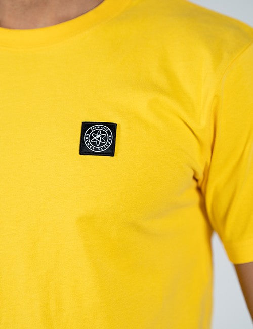 A close-up of a bright yellow short-sleeve t-shirt worn by a model, featuring a black square patch Rocket Science logo on the left chest.
