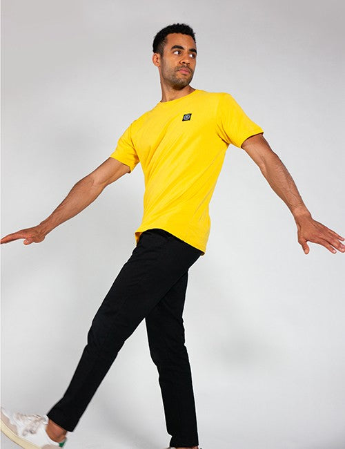 A man wearing a bright yellow short-sleeve t-shirt with a small black Rocket Science logo on the left chest and black pants, posing dynamically against a light gray studio background.