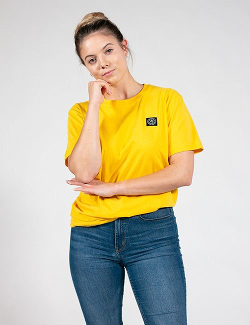 A woman wearing a bright yellow short-sleeve crewneck t-shirt with a small black square Rocket Science logo patch on the left chest, paired with blue jeans, photographed against a light gray studio background.