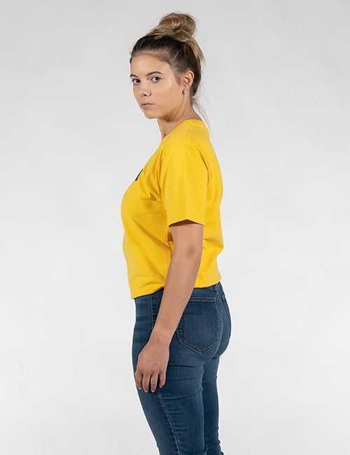 A woman with her hair in a bun is shown from the side profile, wearing a bright yellow short-sleeve t-shirt and dark blue jeans against a plain light gray background.