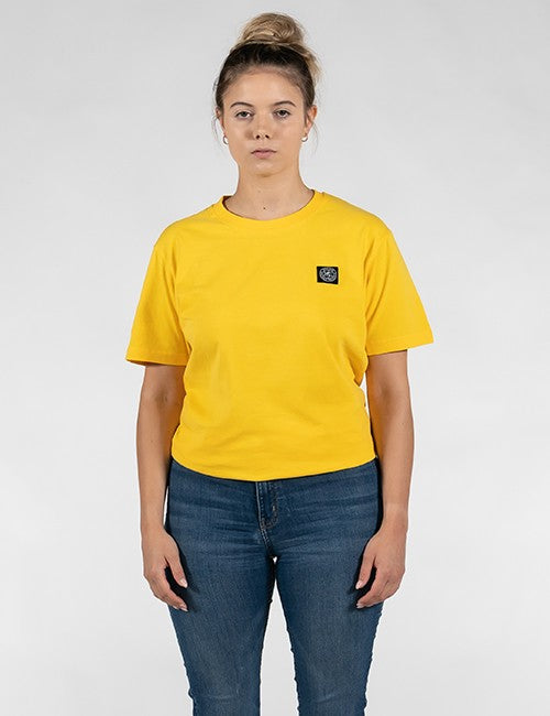 A woman modeling a bright yellow short-sleeve crewneck t-shirt with a small black square Rocket Science logo patch on the left chest, paired with blue jeans, against a light gray studio background.
