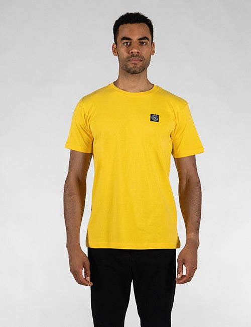 A man wearing a bright yellow crew-neck t-shirt with a small black square Rocket Science logo patch on the left chest, paired with black pants, photographed against a neutral light gray background.