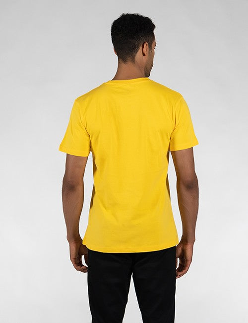 A man viewed from behind wearing a bright yellow short-sleeve crewneck t-shirt and black pants, standing against a light gray studio background.