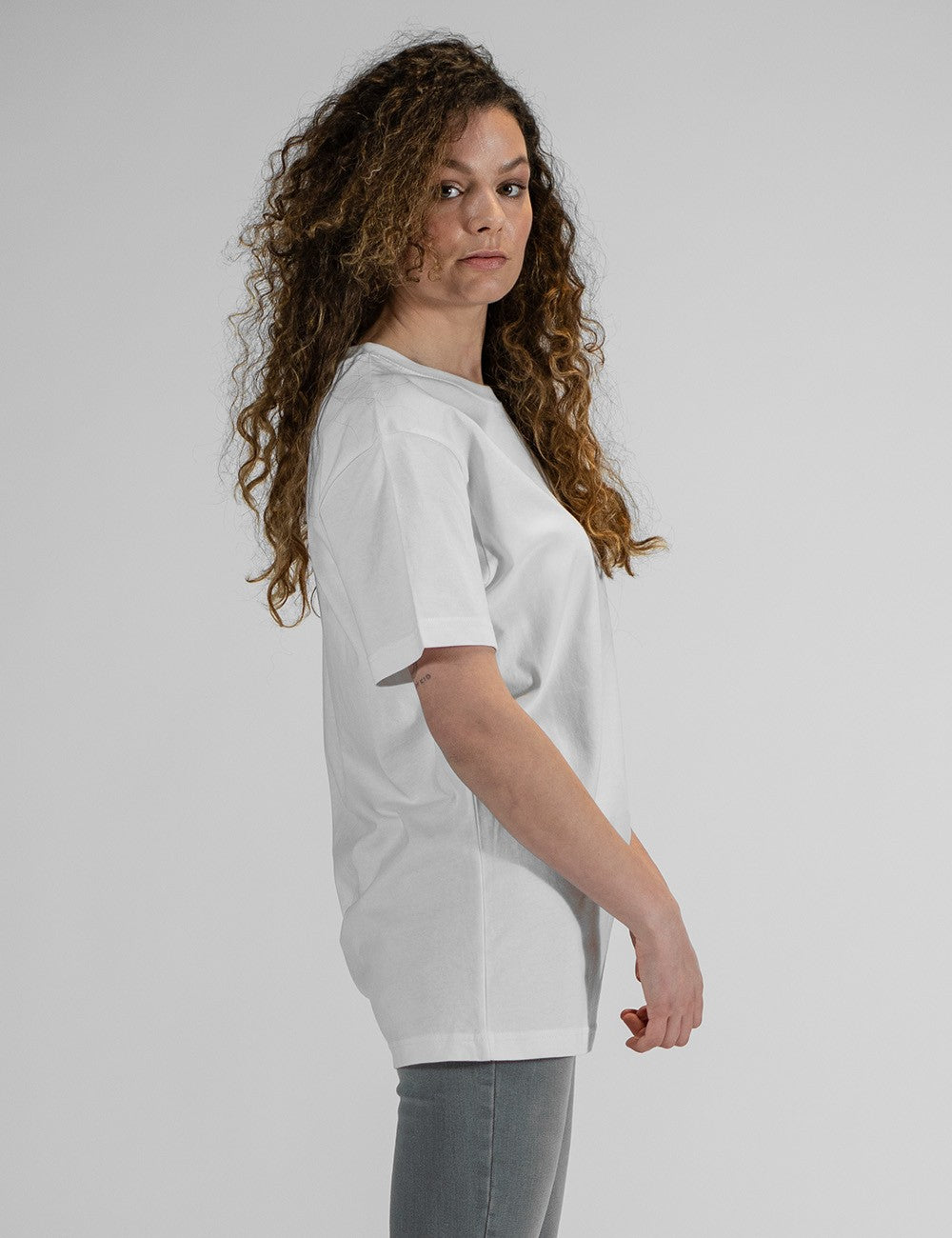 Side profile of a young woman with long, curly brown hair wearing an oversized plain white t-shirt and grey pants against a light grey background.