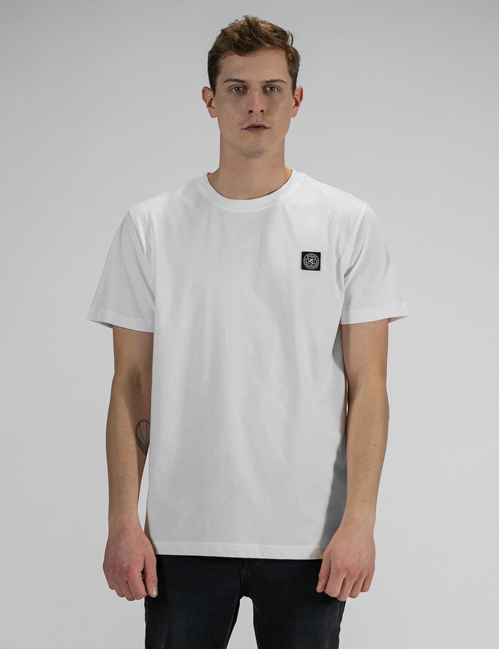 A front view of a young man with short brown hair, wearing a white crewneck t-shirt with a small square black and white brand logo patch on the left chest, and dark jeans.