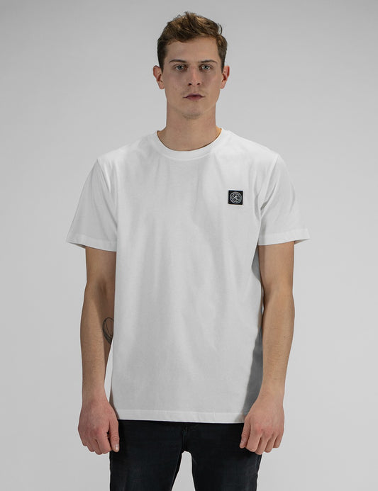 A front view of a young man with short brown hair, wearing a white crewneck t-shirt with a small square black and white brand logo patch on the left chest, and dark jeans.