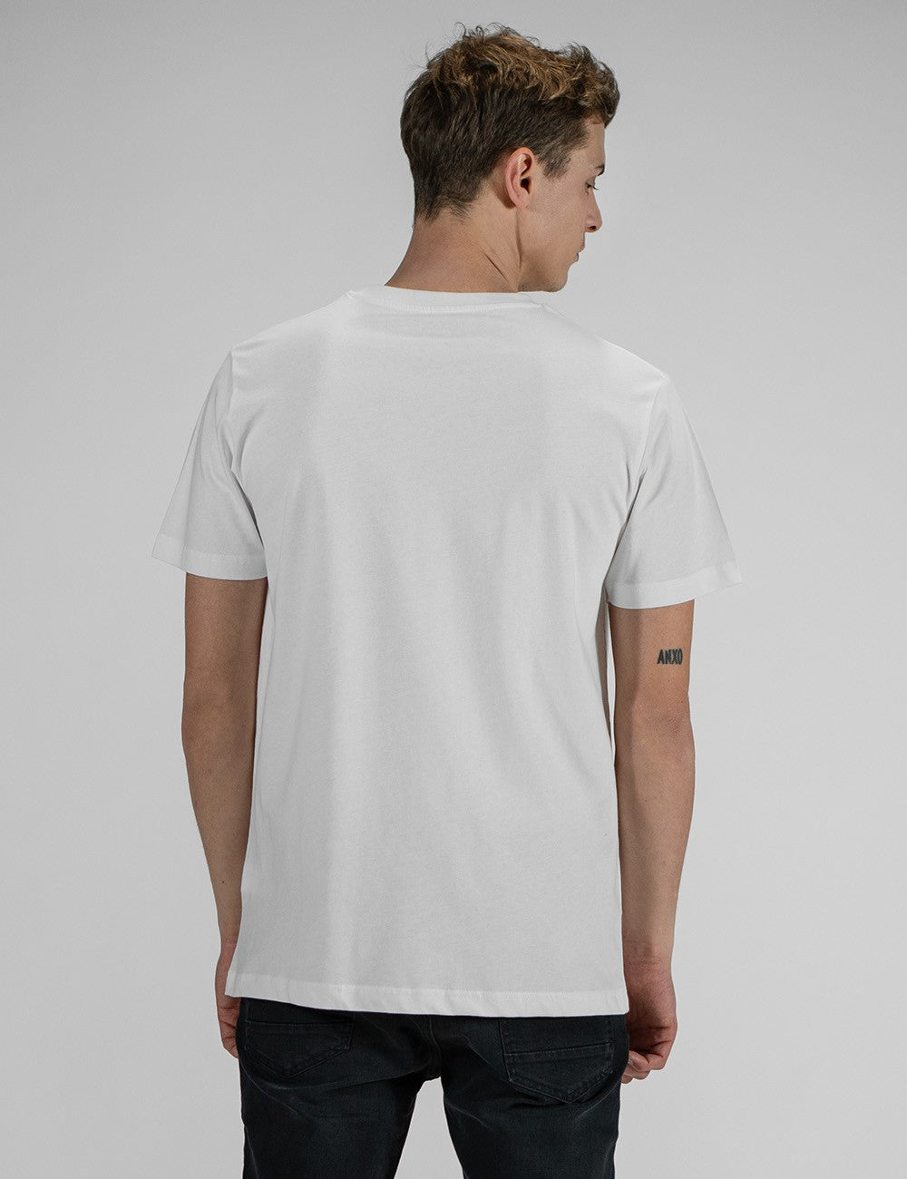 A rear angle image of a young man, wearing a plain white t-shirt and black pants, looking back over his shoulder.